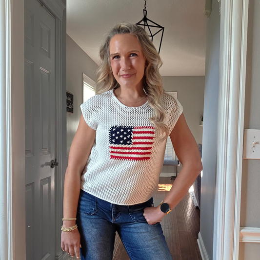 Woman wearing a white crochet top with an American flag design and blue jeans indoors.